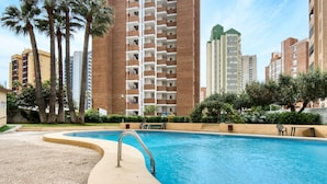 A swimming pool with a metal ladder, surrounded by a landscaped area with palm trees and a brick building in the background.
