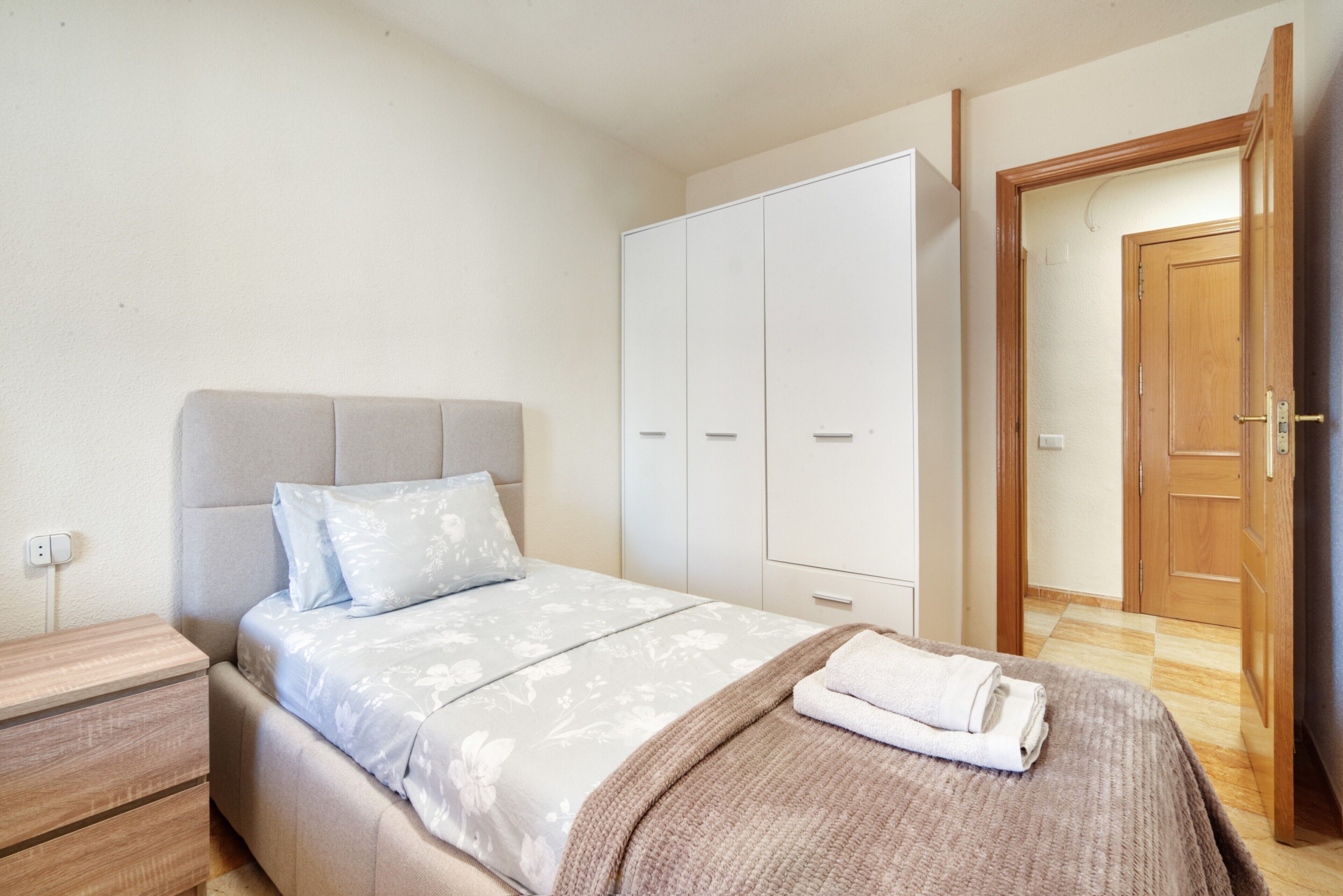 A bedroom with a bed, a wardrobe, a nightstand, and a door leading to another room.