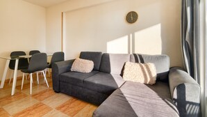 A modern living room with a grey sectional sofa, two armchairs, a glass dining table with chairs, and a clock on the wall.