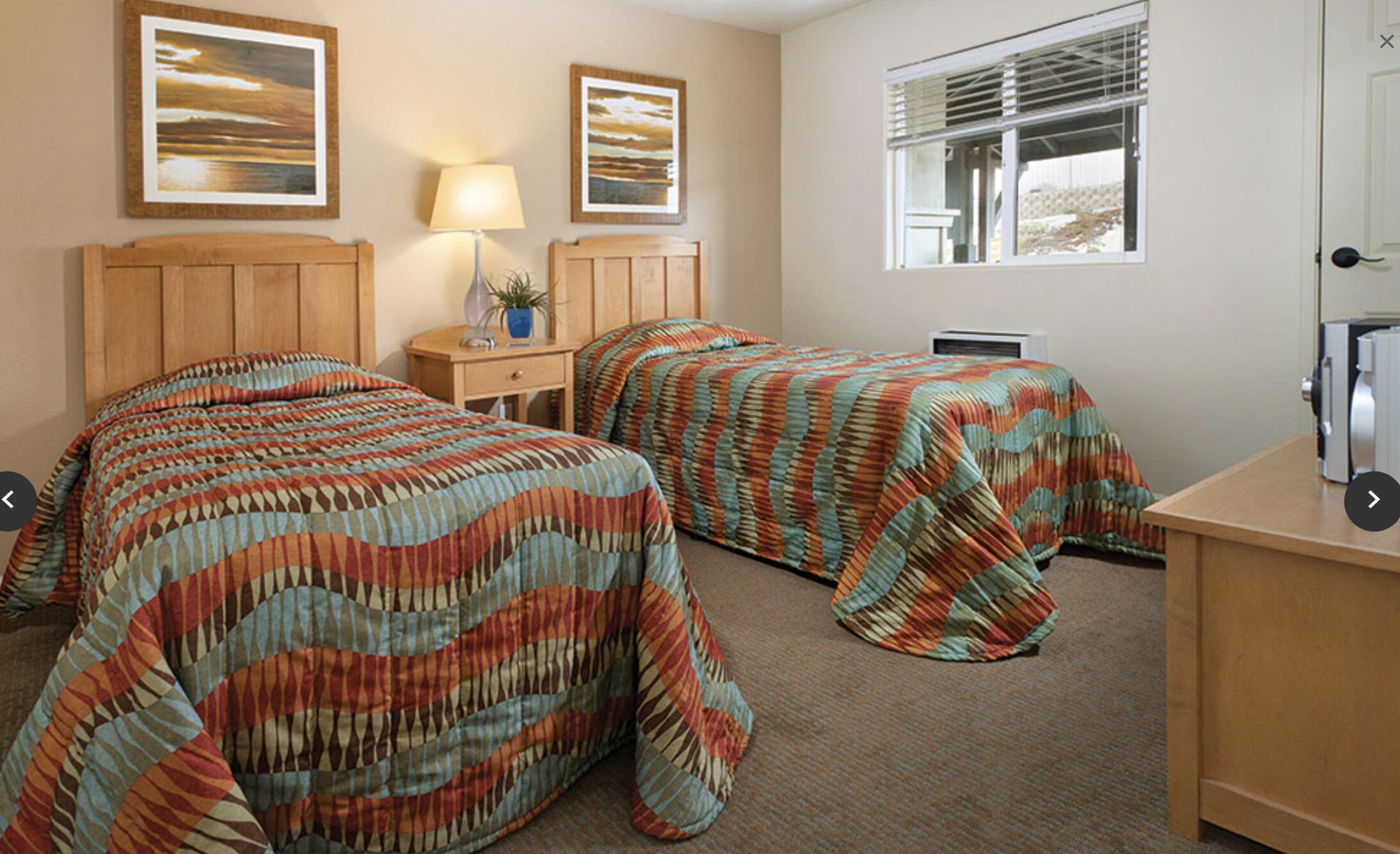 Quarto de hotel com duas camas, uma mesa, uma luminária e uma janela com persianas.