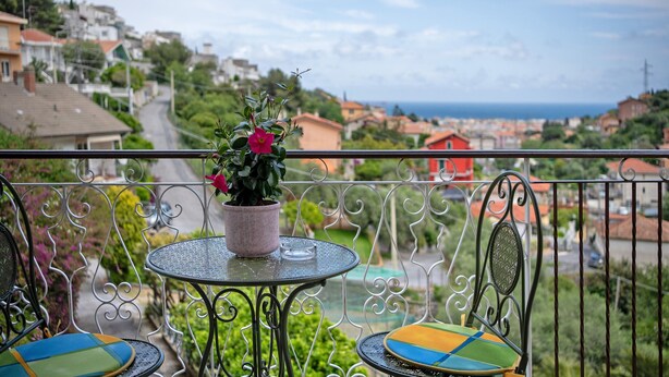 A balcony with a table and chairs, a potted plant, and a view of a coastal town.