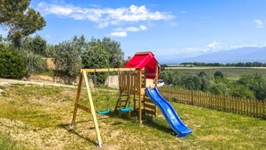Une aire de jeux avec des balançoires et un toboggan, entourée d’une clôture en bois et d’une pelouse.