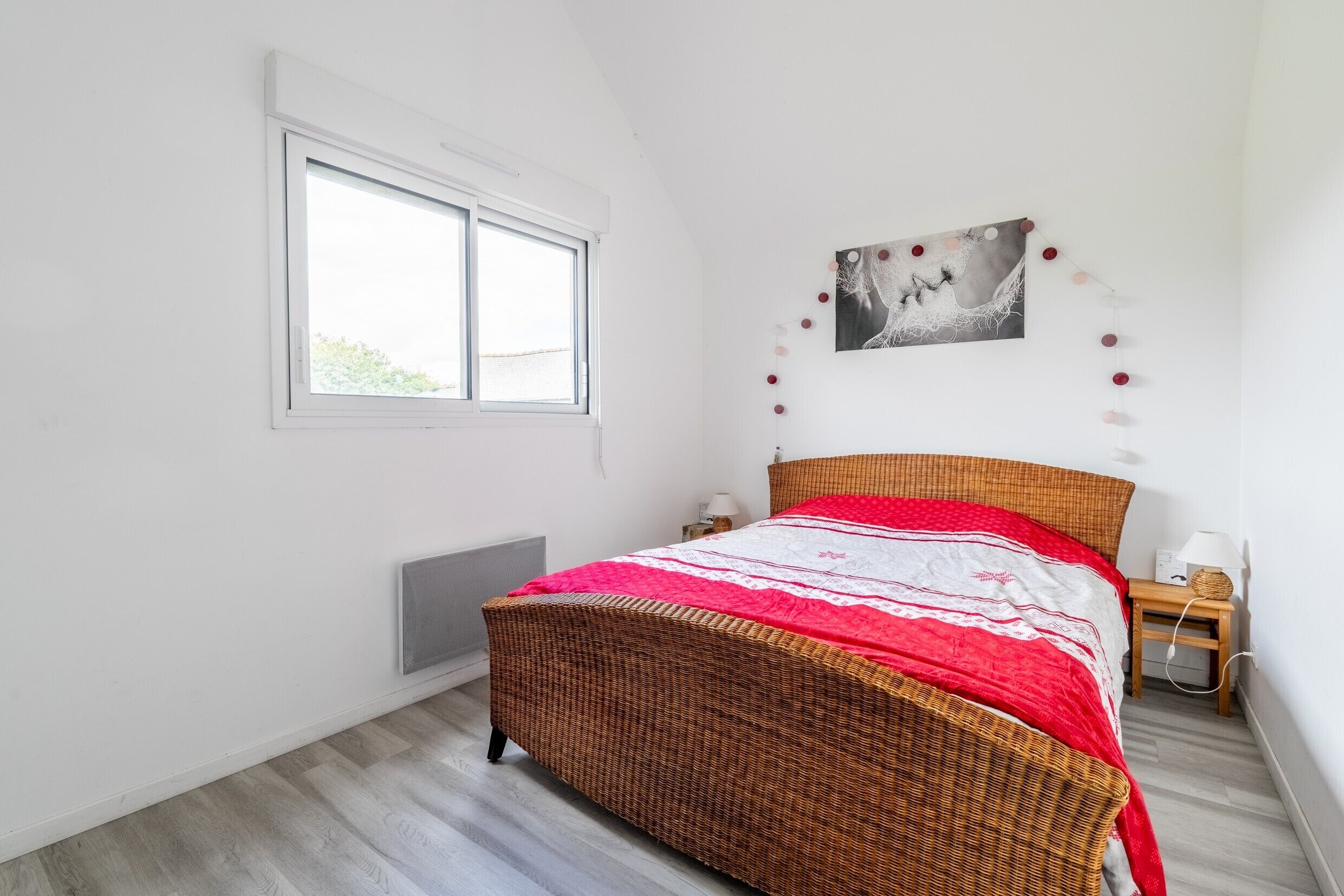 Um quarto com uma cama de vime, uma janela, um quadro na parede e uma mesa de cabeceira com abajur.
