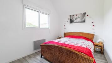 Um quarto com uma cama de vime, uma janela, um quadro na parede e uma mesa de cabeceira com abajur.