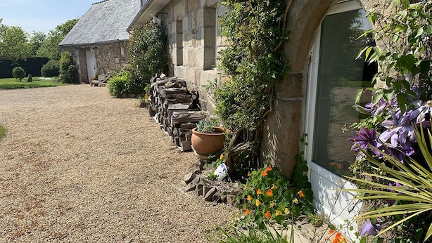 Une construction en pierre avec une allée de gravier, une pile de bois de chauffage et une grande plante en pot.