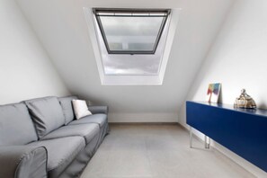 A room with a sloped ceiling, a skylight, a grey sofa, a blue console table, and a tiled floor.