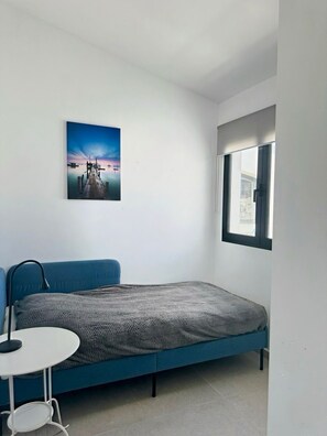 A bedroom with a bed, a small table, a chair, a window, and a wall-mounted picture.