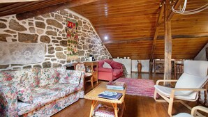 A cozy attic room with a stone wall, wooden ceiling, a floral sofa, a red chair, a wicker coffee table, and a white armchair. - Room at 'Casa Do Largo - First Floor' with Shared Terrace, Shared Garden, and Wi-Fi (Mendiga)