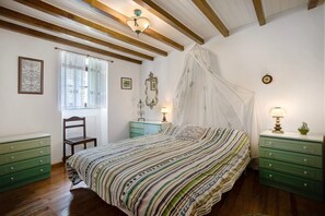 Ein Schlafzimmer mit Bett, Stuhl, Nachttisch, Lampe, einem Fenster mit Vorhängen, einer Wanduhr und einem Deckenventilator.