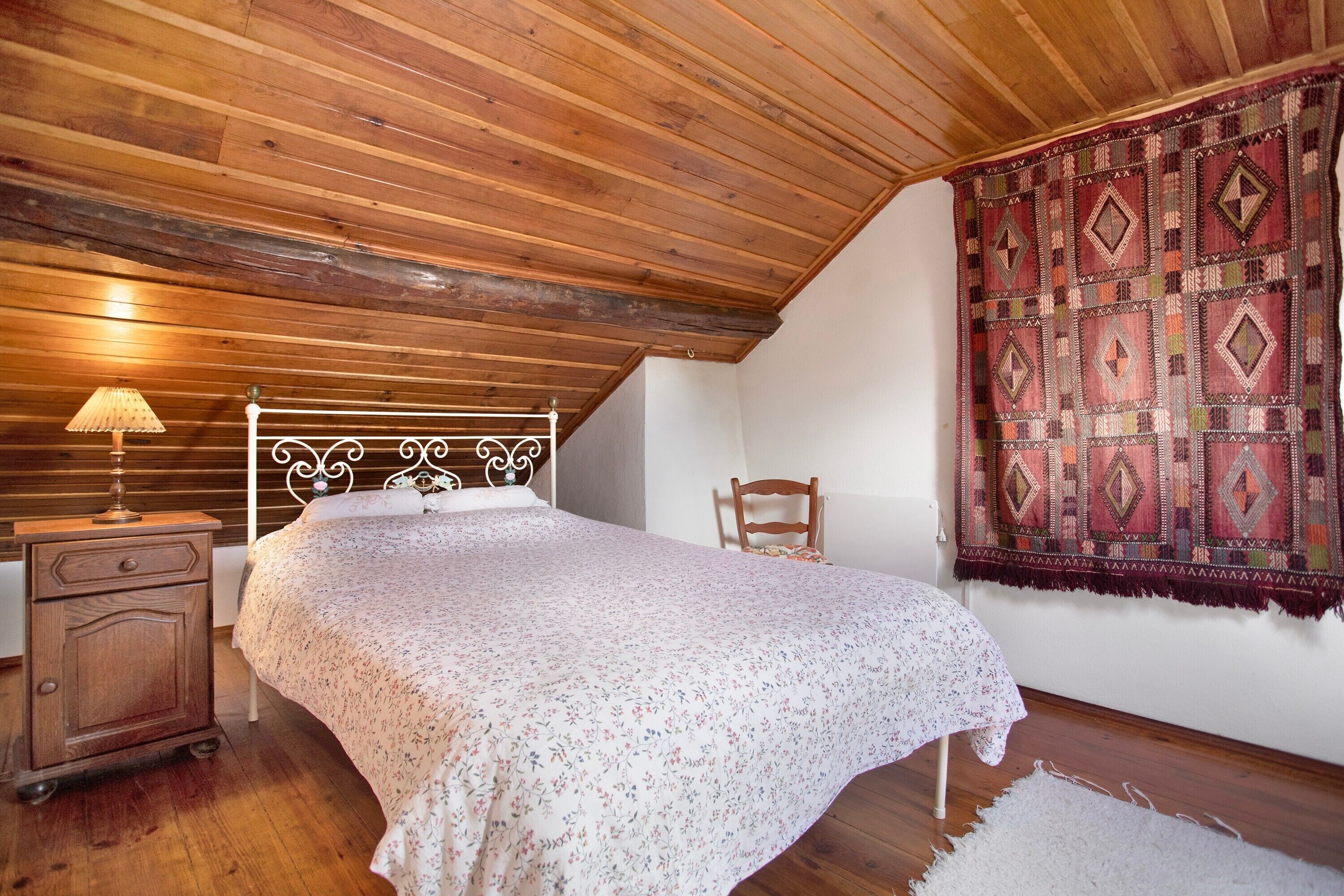 A bedroom with a wooden ceiling, a bed with a floral bedspread, a nightstand, a chair, and a patterned wall hanging.