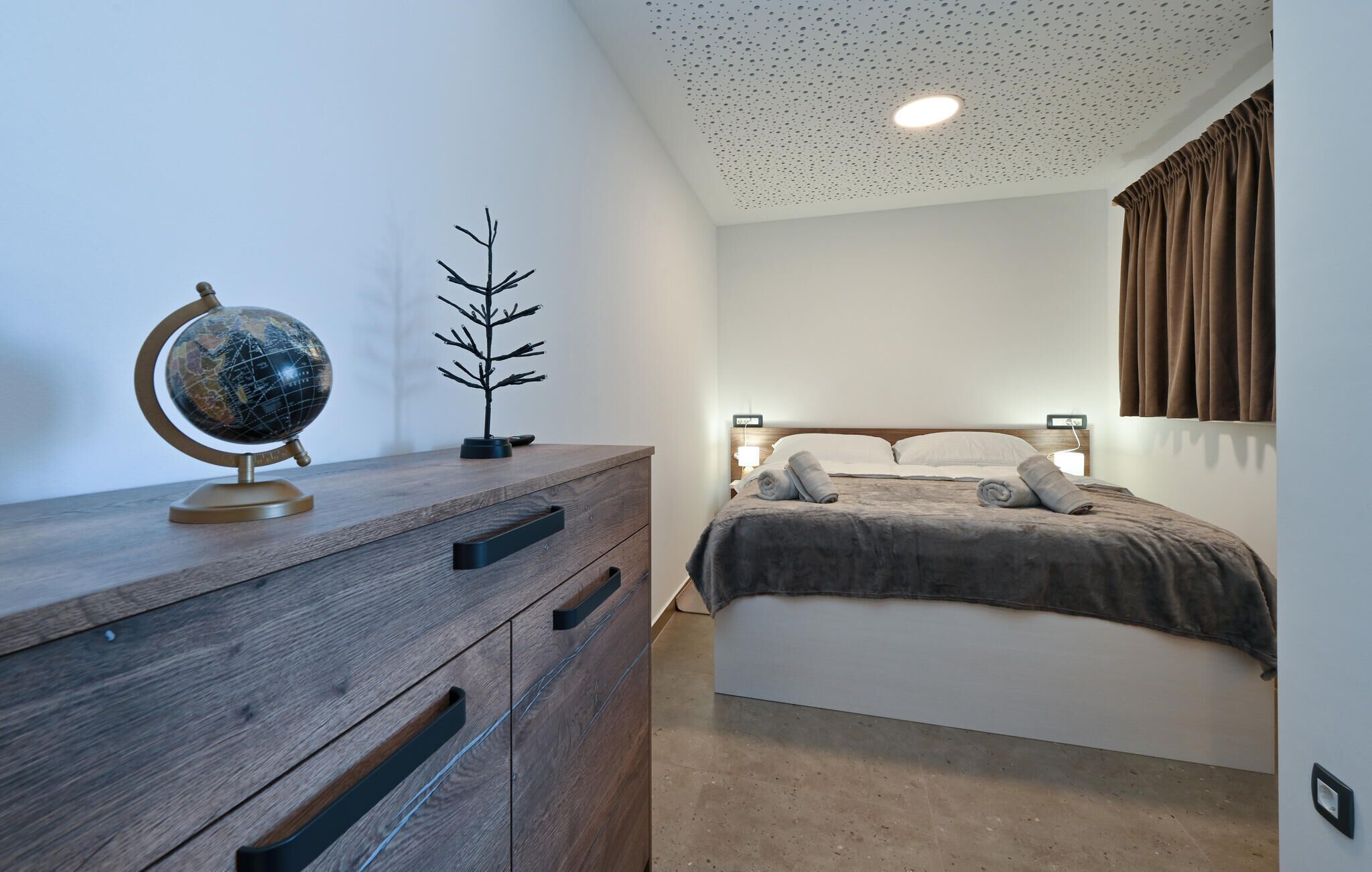 A bedroom with a wooden dresser, a globe, a small tree model, a bed with a headboard, and a ceiling with a light fixture.