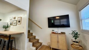 A modern living space with a staircase, a dining area with bar stools, a television mounted on the wall, and wicker furniture. - The Birch at Lazy Oaks Resort (duplex) (Lakeside)