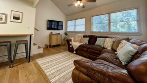 A living room with a brown leather couch, a wooden coffee table, a TV mounted on the wall, and a ceiling fan. - The Birch at Lazy Oaks Resort (duplex) (Lakeside)