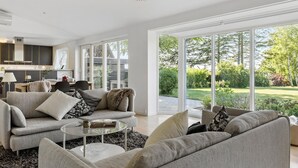 Un salón espacioso con un gran ventanal que da a un jardín, una cocina con muebles oscuros y una lámpara moderna.