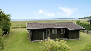 A small, dark wooden building with a sloped roof, a single door, and a window. It is situated on a grassy field with a view of the sea in the background.