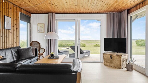 A modern living room with a wooden ceiling, a black leather sofa, a dining table, a TV, and a large sliding glass door leading to an outdoor area with a view of the landscape.