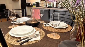 A modern dining area with a dark wood table set for four, including plates, cutlery, and wine glasses. A vase with purple flowers is on the table, and there is a kitchen in the background.