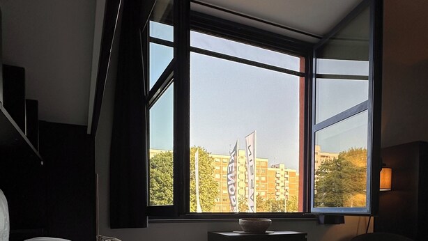 A hotel room with a large bed, a view of buildings through the window, and a small table with a bowl on it.