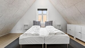 A bedroom with a sloped ceiling, a window, two beds with white bedding, patterned pillows, a bedside table, a lamp, and a wardrobe.