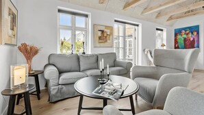 A modern living room with a grey sofa, a round coffee table, and a grey armchair. There are two windows with white frames, a painting on the wall, and a vase with dried flowers on a side table.