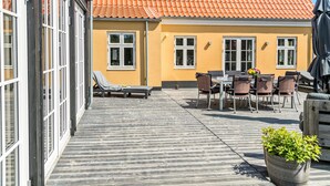 A covered outdoor area with a wooden deck, lounge chairs, and a table with chairs.