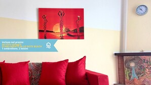 Een rode bank met gekleurde kussens, een wandklok en een schilderij van twee personen in de zonsondergang.