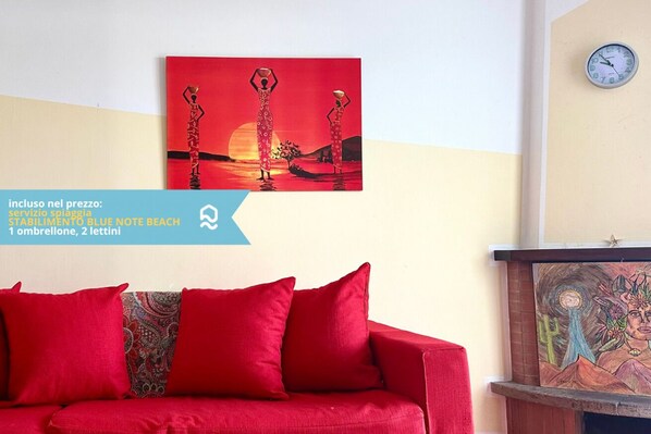 A red sofa with patterned cushions, a wall-mounted clock, and a painting of two figures with a sunset.