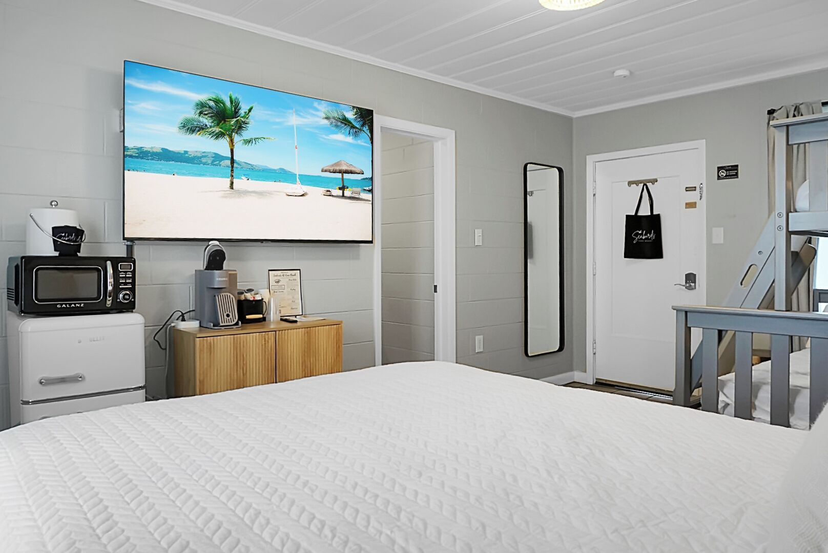 A bedroom with a large bed, a television displaying a beach scene, a microwave, a coffee maker, and a wooden cabinet.
