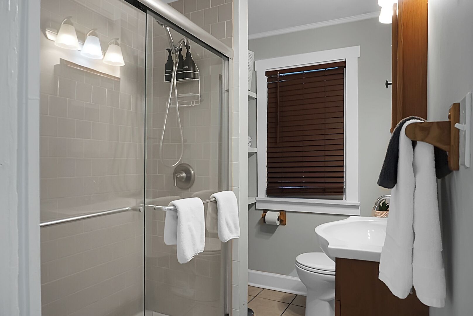 Un baño moderno con ducha envuelta en vidrio, un lavamanos blanco y una ventana con persianas.