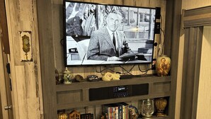 Ett vardagsrum med en platt-TV som är monterad på en väggpanel av trä, en öppen spis nedanför och en bokhylla med diverse föremål.