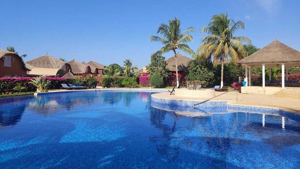 A clear blue swimming pool surrounded by thatched-roof huts and palm trees, with lounge chairs and a gazebo in the background. - 2 Bedroom Villa in Saly (Saly)