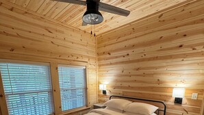 Un dormitorio de cabaña de madera con una cama, dos mesitas de noche, un ventilador de techo y una ventana con persianas.