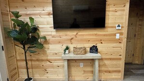 Pared de madera con un televisor de pantalla plana montado sobre una pequeña mesa que sostiene una planta en maceta, una cesta tejida y un letrero.