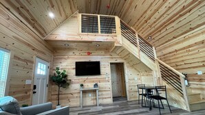 Una cabaña de madera espaciosa con un dormitorio en el altillo, comedor y sala de estar con sofá y televisor.