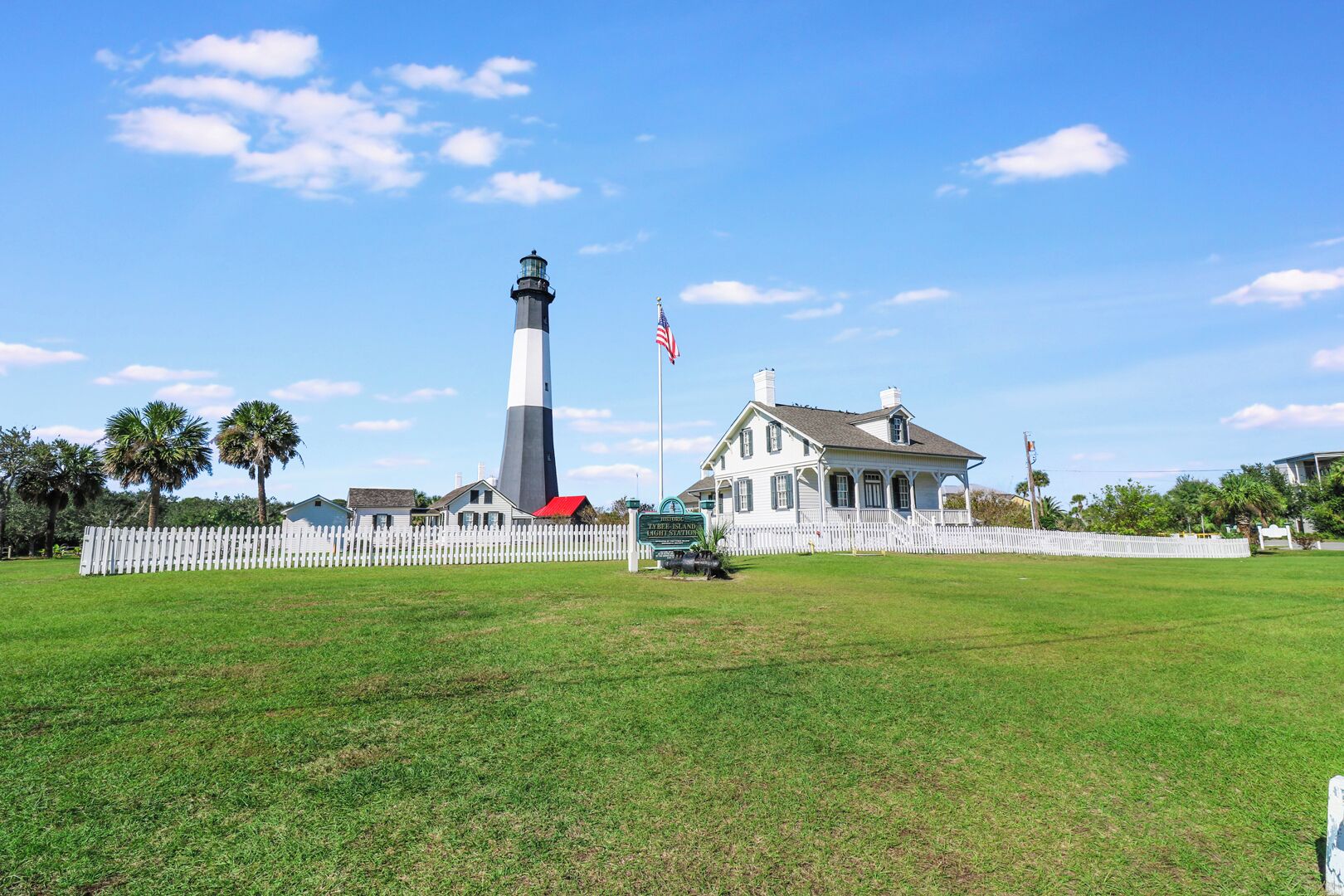 A historic lighthouse with a white and black tower, a white picket fence, and a large white building with a red roof in the foreground.