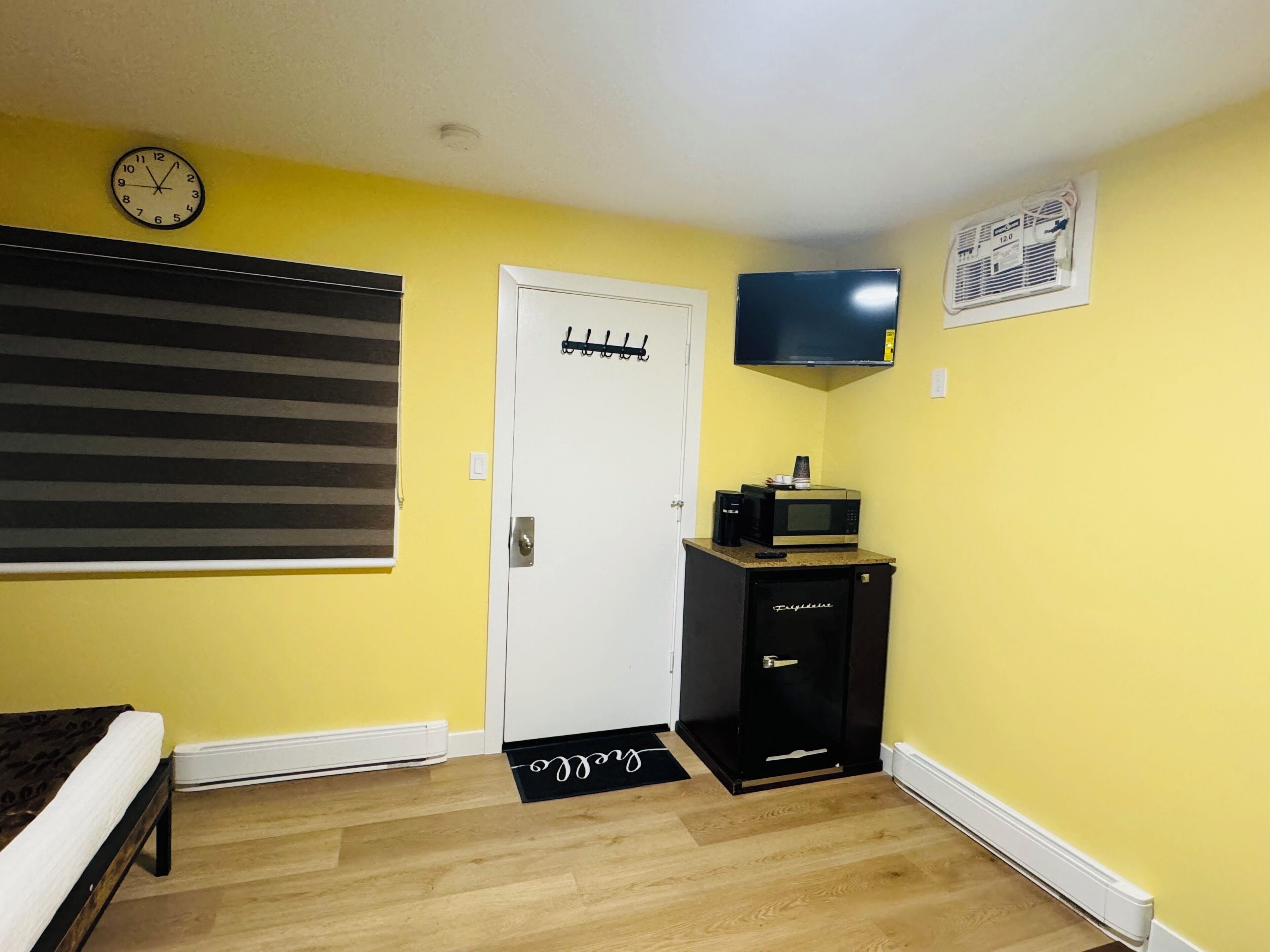 Una habitación con paredes amarillas, una puerta blanca, un armario negro con microondas, un reloj en la pared y una persiana ventana a rayas.