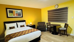 Una habitación de hotel con una cama, una silla, una mesita, un reloj y una ventana con persianas.