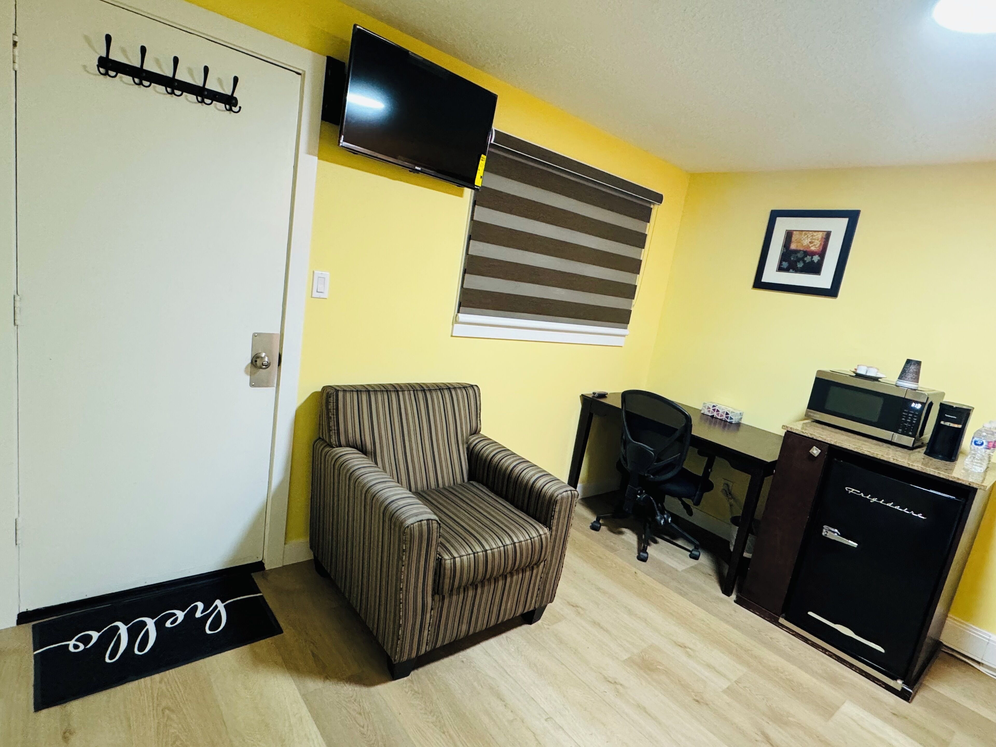 Una habitación compacta con un sillón a rayas, un escritorio con computadora y microondas, y una kitchenette pequeña.