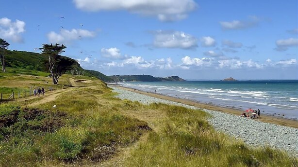 Un paysage côtier avec un sentier herbeux menant à une plage, quelques personnes qui se promènent et une île visible au loin.