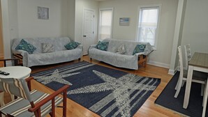 Una sala de estar con dos sofás grises, un tapete azul y blanco estampado, una mesita blanca y una mesa de comedor de madera con sillas.