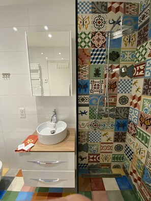 Ein Badezimmer mit einem weißen Waschbecken, einem Spiegel, einer Dusche mit Duschbrause und einer gefliesten Wand mit verschiedenen Mustern.
