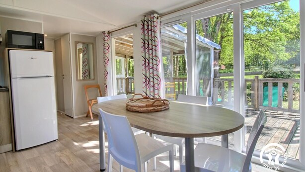 Una cocina moderna con microondas y refrigerador integrados, una mesa de comedor redonda con sillas blancas y un amplio ventanal corredizo que da a una terraza con vistas a árboles.