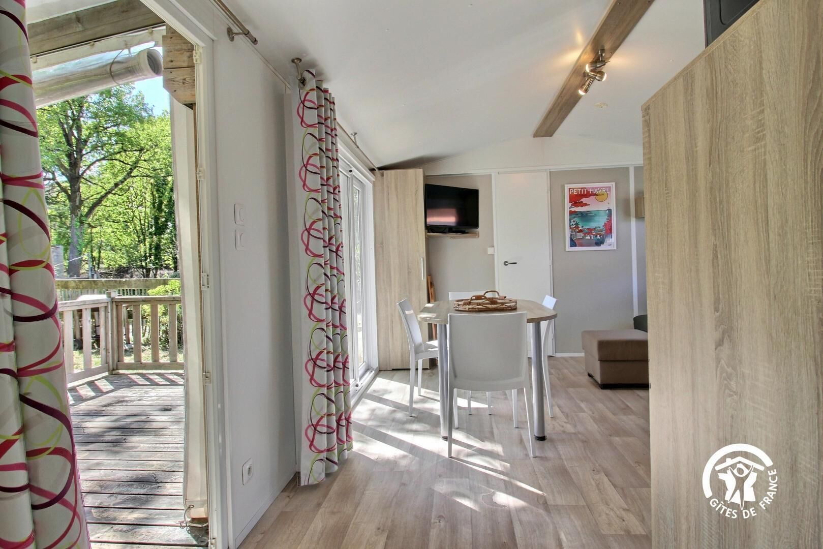 A modern interior with a wooden floor, a dining area with a table and chairs, a TV mounted on the wall, and a view of the outdoors through a glass door.