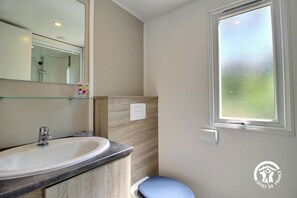Een badkamer met een wit wastafel, een spiegel, een glazen plank, een raam en een lichtschakelaar aan de muur.