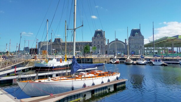 Un puerto deportivo con barcos amarrados, un edificio grande con una torre de reloj y una zona cubierta con gente.