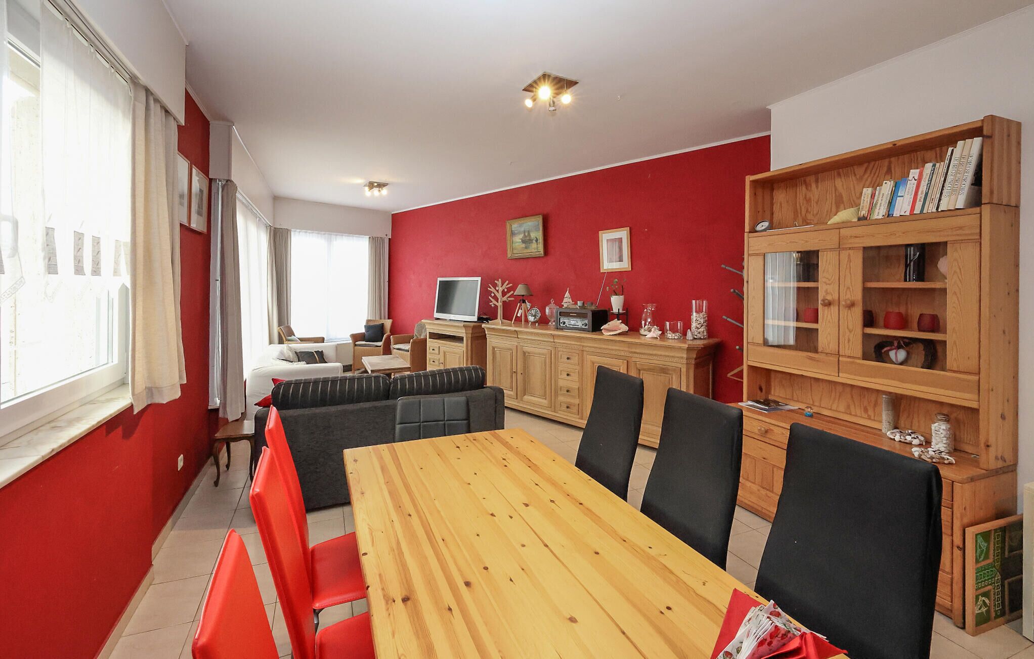 A dining area with a wooden table and red chairs, a television, and a bookshelf with various items.