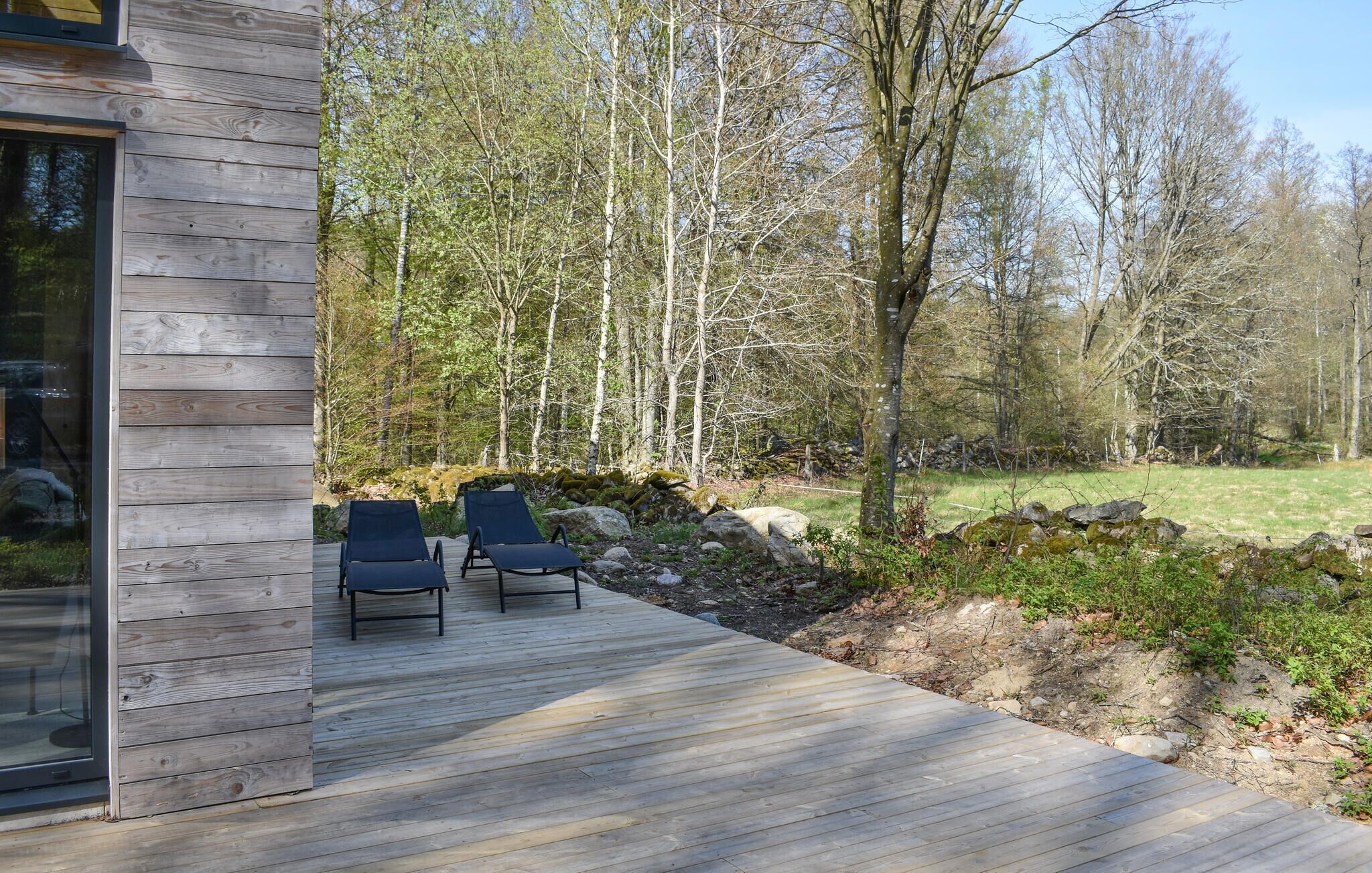A wooden deck with two lounge chairs, a stone wall, and a forested area in the background.