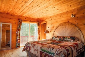 Un dormitorio de cabaña de madera con una cama grande, una mesita de noche, una lámpara y una ventana con cortinas.