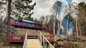 Puinen terassi johtaa punaiseen mökkiin, jossa on parveke. Mökkiä ympäröivät puut ja kivinen maisema.