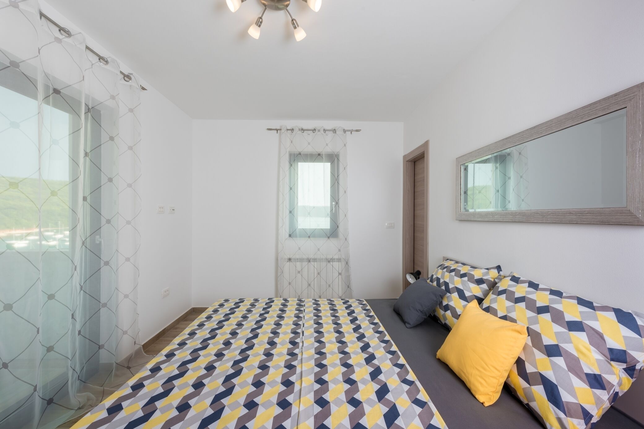 A modern bedroom with a geometric-patterned bedspread, a large mirror, and a view of the outdoors through a window with a geometric curtain.